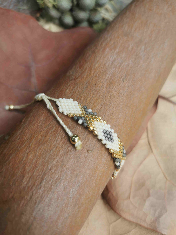 Bracelet artisanal colombien tissé à la main avec des perles dorées, blanches et grises, posé sur un fond naturel.