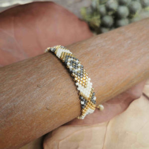 Bracelet artisanal colombien tissé à la main avec des perles dorées, grises et blanches, posé sur un fond naturel.