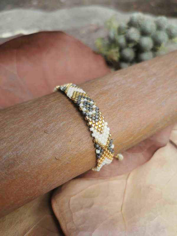 Bracelet artisanal colombien tissé à la main avec des perles dorées, grises et blanches, posé sur un fond naturel.