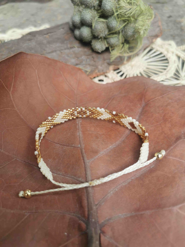 Bracelet artisanal colombien tissé à la main avec des perles dorées, marrons et blanches, posé sur un fond naturel.