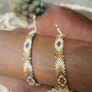 Une paire de bracelets artisanaux de Colombie tissée à la main avec des perles dorées, blanches, grises et marrons posée sur un fond naturel.